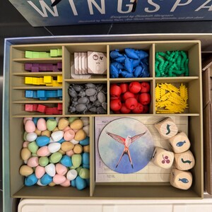 May include: A board game organizer with various components. The organizer has multiple compartments filled with colorful game pieces, including blue, red, green, and yellow shapes, along with dice and tokens. The game box lid reads "Wingspan".
