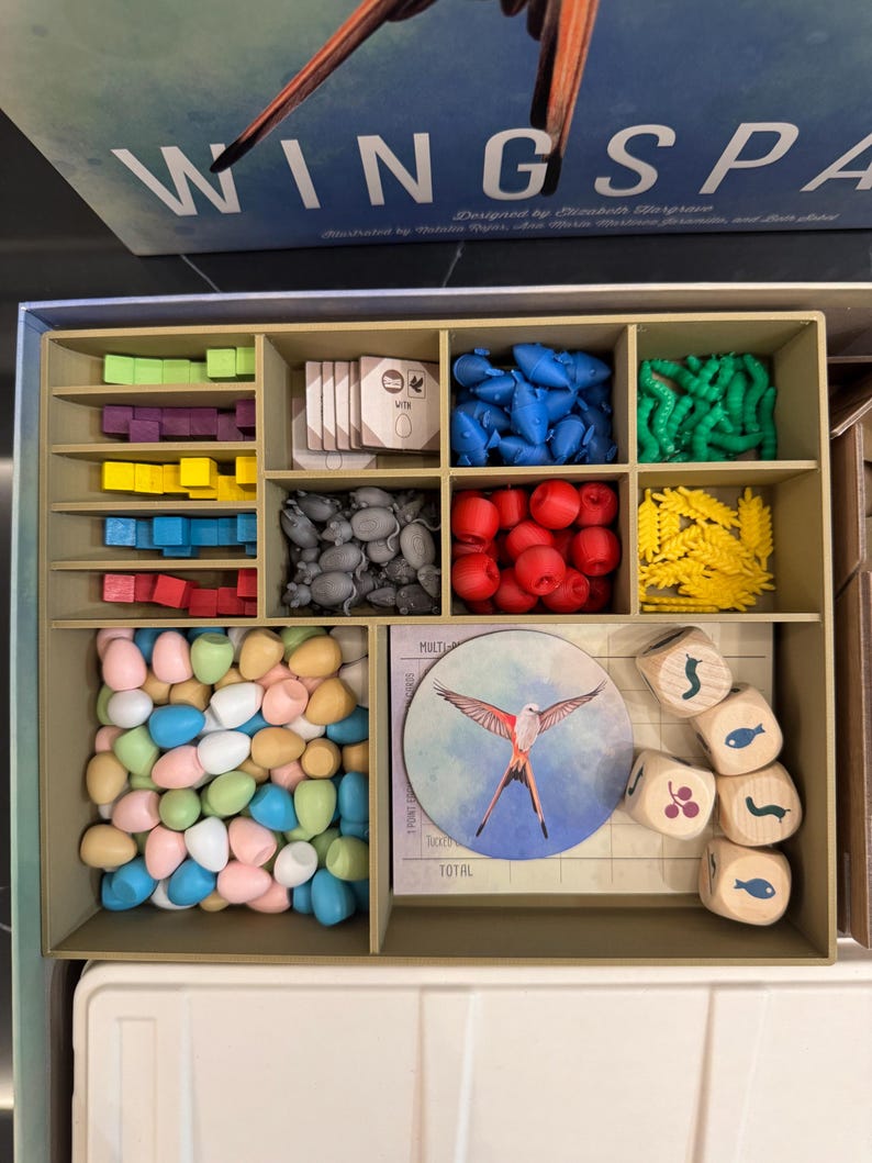May include: A board game organizer with various components. The organizer has multiple compartments filled with colorful game pieces, including blue, red, green, and yellow shapes, along with dice and tokens. The game box lid reads "Wingspan".