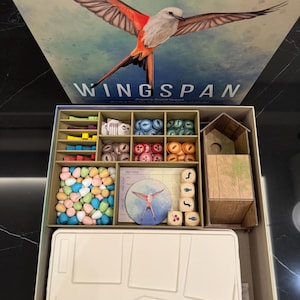 May include: A board game box titled "Wingspan" featuring a bird illustration. The open box reveals game components: wooden dice, colorful tokens, and a miniature birdhouse. The box is beige and the game components are in a variety of colors.
