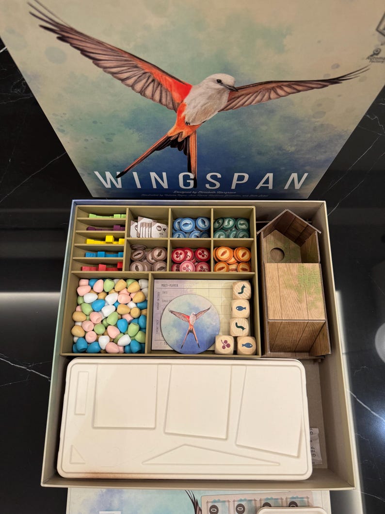 May include: A board game box titled "Wingspan" featuring a bird illustration. The open box reveals game components: wooden dice, colorful tokens, and a miniature birdhouse. The box is beige and the game components are in a variety of colors.