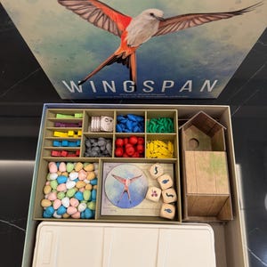 May include: The board game "Wingspan" is displayed open, revealing game pieces and components. The box art features a bird with outstretched wings. Inside, there are various colored game pieces, including eggs, dice, and wooden tokens. The game box is beige and the game pieces are in a tray.