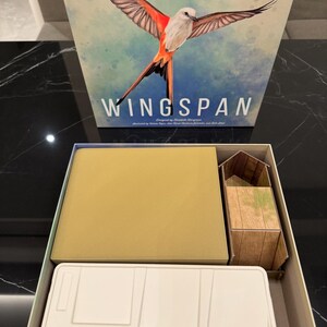 May include: A board game box with the title "Wingspan" and an illustration of a bird with orange and brown wings. The box is open, revealing game components, including a tan insert and a small wooden-looking structure.