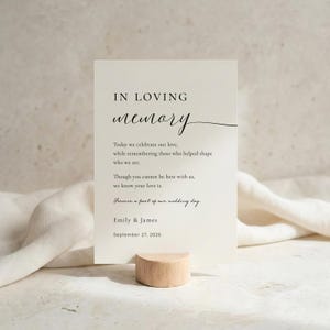 May include: A rectangular cream-colored memorial sign featuring black text that reads: IN LOVING memory, Today we celebrate our love, while remembering those who helped shape who we are. Though you cannot be here with us, we know your love is. Forever a part of our wedding day. Emily & James, September 27, 2026. The sign is held upright by a small, cylindrical wooden stand.