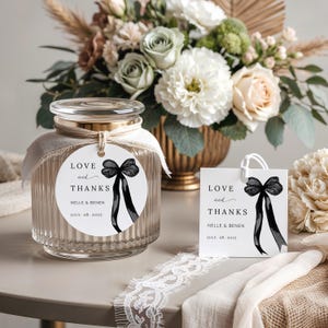 May include: A clear glass jar with a lid and a round tag that says "LOVE and THANKS" with a black bow illustration. A matching gift tag is also present. The jar is tied with a ribbon and sits on a table with a floral arrangement.