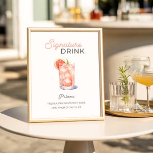 以下が含まれることがあります： 「Signature Drink」の文字と、パロマカクテルの水彩イラストが描かれた額入りのサイン。材料は飲み物の画像の下に記載されています。サインは白いテーブルの上にあります。