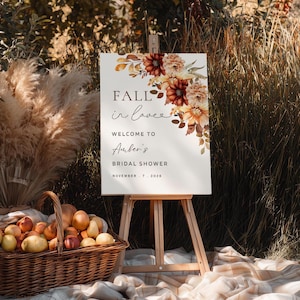Puede incluir: Un letrero blanco sobre un caballete de madera con el texto "Fall in love" y "Welcome to Amber's Bridal Shower". El letrero está decorado con arreglos florales en tonos naranja, marrón y amarillo. Una cesta de fruta y hierba de la pampa están en primer plano.