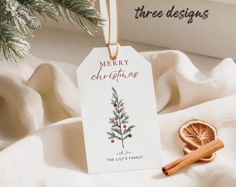 Könnte beinhalten: Weißes Geschenk-Etikett mit einer Weihnachtsbaum-Illustration, roten Akzenten und dem Text "Merry Christmas". Ein cremefarbenes Band ist befestigt. Das Etikett liegt auf einer weißen Oberfläche mit einem Tannenzweig und getrockneten Orangenscheiben.