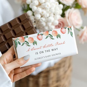 May include: A rectangular chocolate bar wrapped in white paper featuring a watercolor peach and leaf illustration. The text reads A Sweet Little Peach IS ON THE WAY and AUDREY'S BABY SHOWER in a mix of script and sans-serif fonts.