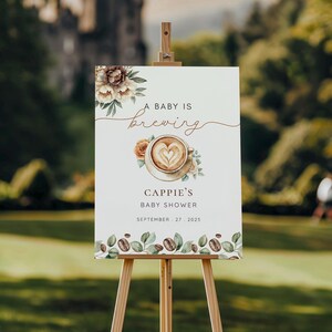 May include: A white sign on a wooden easel with the text "A Baby Is Brewing" and "Cappie's Baby Shower, September 27, 2025". The sign features a coffee cup with a heart design and floral accents, with coffee beans and leaves at the bottom.