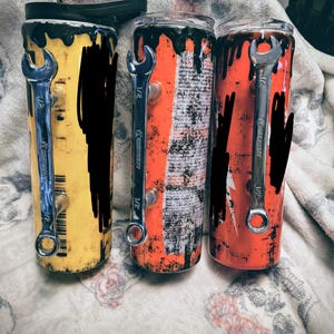 May include: Three stainless steel tumblers with black and yellow, orange, and red designs. Each tumbler has a wrench attached and a black lid. The tumblers are cylindrical and appear to be insulated.