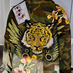 May include: A camouflage jacket featuring a large embroidered tiger head with golden wings, floral patches, and a white rectangular fabric label with the text MORE AMOR POR FAVOR stitched on the upper left side of the back panel.