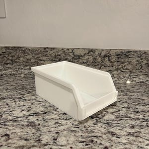 May include: A white rectangular storage bin with a sloped front. The bin is made of a smooth, solid material and sits on a speckled countertop. The bin is designed for organizing items.