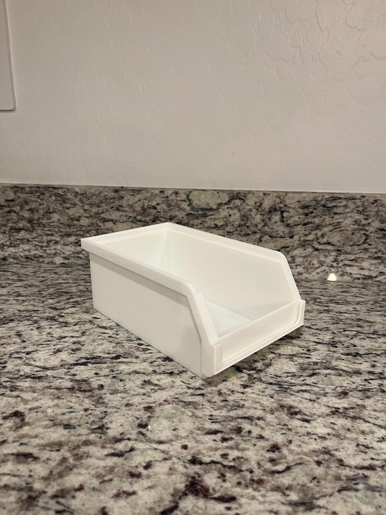 May include: A white rectangular storage bin with a sloped front. The bin is made of a smooth, solid material and sits on a speckled countertop. The bin is designed for organizing items.