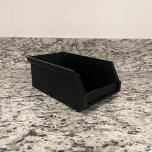 May include: A black rectangular storage bin with a sloped front and a flat base. The bin is made of a solid material and is sitting on a speckled countertop. The bin is empty.