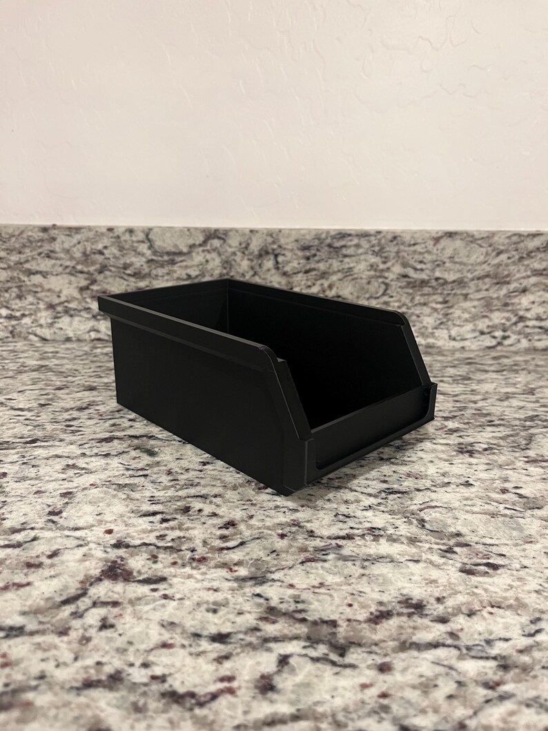 May include: A black rectangular storage bin with a sloped front and a flat base. The bin is made of a solid material and is sitting on a speckled countertop. The bin is empty.