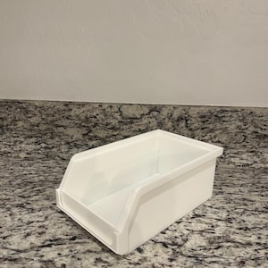 Puede incluir: Un contenedor de almacenamiento rectangular blanco con un frente inclinado y un recorte en forma de asa. El contenedor está hecho de un material liso y sólido y se encuentra sobre una encimera moteada. Para organizar artículos.