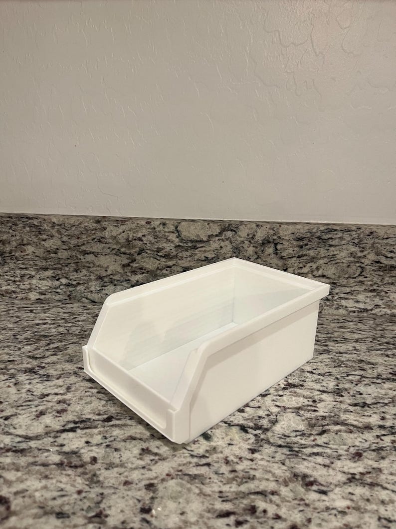May include: A white rectangular storage bin with a sloped front and a handle-like cutout. The bin is made of a smooth, solid material and sits on a speckled countertop. The bin is designed for organizing items.