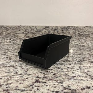 May include: A matte black storage bin with a rectangular shape. The bin has a sloped front for easy access and is sitting on a speckled countertop. The bin is designed for organizing small items.