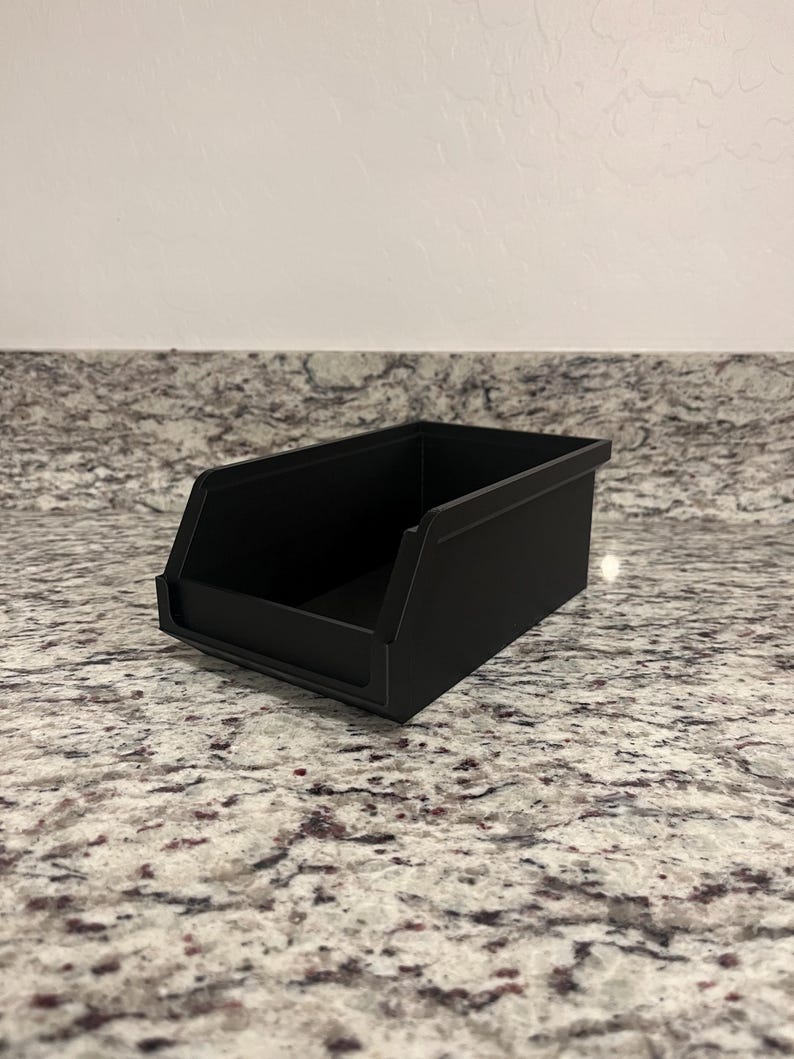 May include: A matte black storage bin with a rectangular shape. The bin has a sloped front for easy access and is sitting on a speckled countertop. The bin is designed for organizing small items.