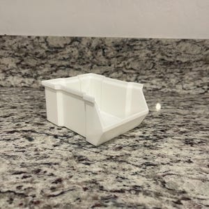 May include: A white plastic storage bin with a rectangular shape and an open front. The bin is designed for organizing small items. The bin is sitting on a granite countertop.
