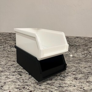 May include: A two-tiered storage bin. The bottom bin is black, and the top bin is white. The bins are rectangular with an open front, designed for easy access to contents. The bins are stacked on top of each other.