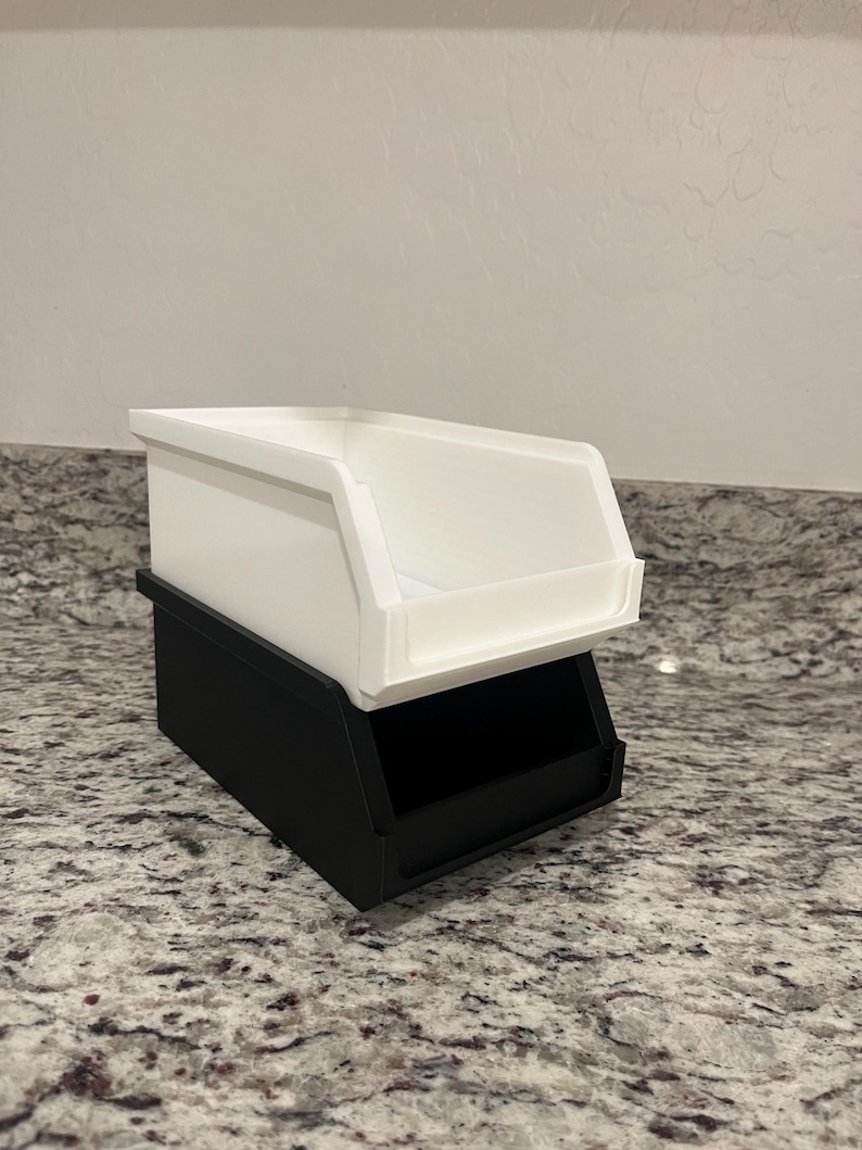 May include: A two-tiered storage bin. The bottom bin is black, and the top bin is white. The bins are rectangular with an open front, designed for easy access to contents. The bins are stacked on top of each other.