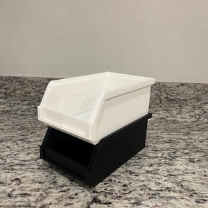 May include: A two-tiered storage bin. The top bin is white, with a sloped front for easy access. The bottom bin is black, providing a stable base. The bins are made of a smooth, matte material and are designed for organizing small items.