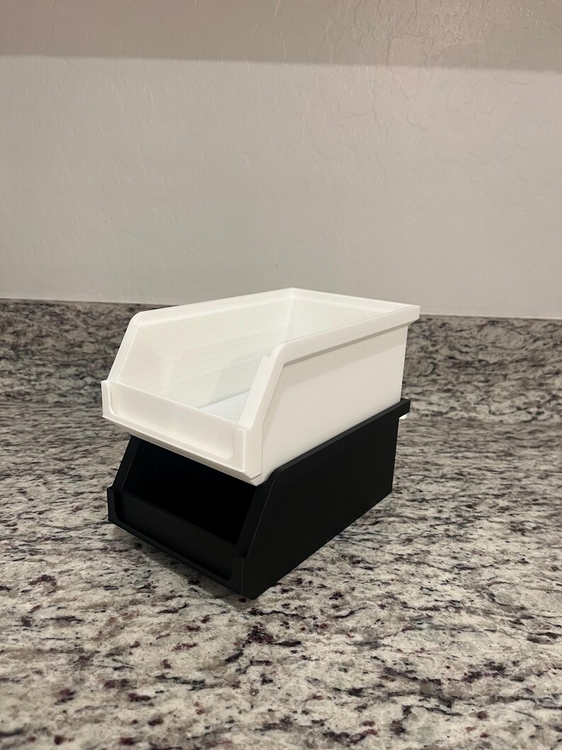 May include: A two-tiered storage bin. The top bin is white, with a sloped front for easy access. The bottom bin is black, providing a stable base. The bins are made of a smooth, matte material and are designed for organizing small items.