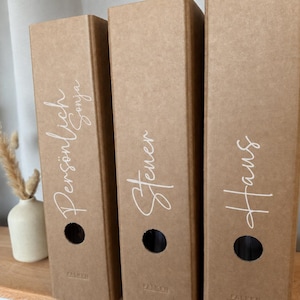 Könnte beinhalten: Drei Aktenordner aus braunem Kraftpapier, die aufrecht stehen. Jeder Ordner hat ein kreisförmiges Griffloch und weiße Beschriftungen in Schreibschrift mit den Wörtern Persönlich Sonja, Steuer und Haus. Der Markenname Falken ist unten eingeprägt.