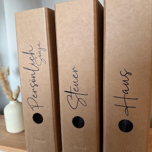 Könnte beinhalten: Drei braune Pappordner mit schwarzen Textetiketten. Die Etiketten lauten "Persönlich Sonja", "Steuer" und "Haus". Jeder Ordner hat ein kreisförmiges Loch und den Markennamen "FALKEN" unten aufgedruckt.