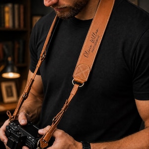 May include: A tan leather camera strap featuring a wide padded section with the text Olivia Willam PHOTOGRAPHY embossed in black. The strap includes adjustable thin leather cords, metal hardware, and reinforced stitching at the connection points.