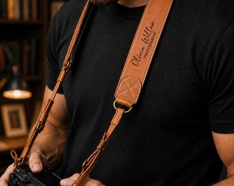May include: A tan leather camera strap featuring a wide padded section with the text Olivia Willam PHOTOGRAPHY embossed in black. The strap includes adjustable thin leather cords, metal hardware, and reinforced stitching at the connection points.