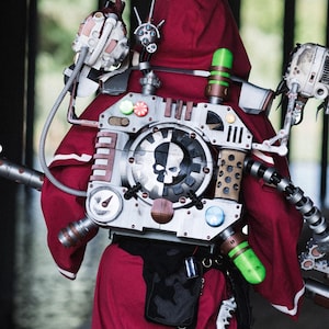 May include: A deep red hooded robe worn with a complex mechanical backpack featuring metallic arms, gears, gauges, and a central circular emblem with a skull icon. The outfit includes black boots and various technical components attached to the back.