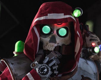 May include: A red hooded robe with white trim and mechanical gear accents, worn with a skull mask featuring glowing green circular eyes. The costume includes robotic shoulder attachments with glowing green lights and various technical components.