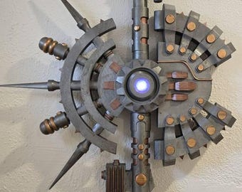 Può includere: Un bastone meccanico con una luce blu centrale, componenti color grigio metallizzato e rame, e punte affilate. Il design include un centro a forma di ingranaggio, una punta superiore e un manico nero testurizzato con dettagli industriali.