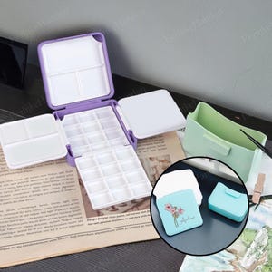 Op de afbeelding: Een paars en wit aquarelpalet met meerdere compartimenten, open om de napjes te laten zien. Een lichtgroene container en een kleine blauwe case met een bloemenontwerp zijn ook zichtbaar. De tekst "Arher" staat op de blauwe case.