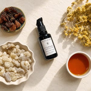 May include: A black pump bottle labeled Flower & Fern Eternal All-in-One Radiance Serum 1 fl oz / 30 ml, surrounded by a small bowl of dark resin, a scalloped bowl of light resin chunks, a small cup of amber liquid, and dried yellow flowers.