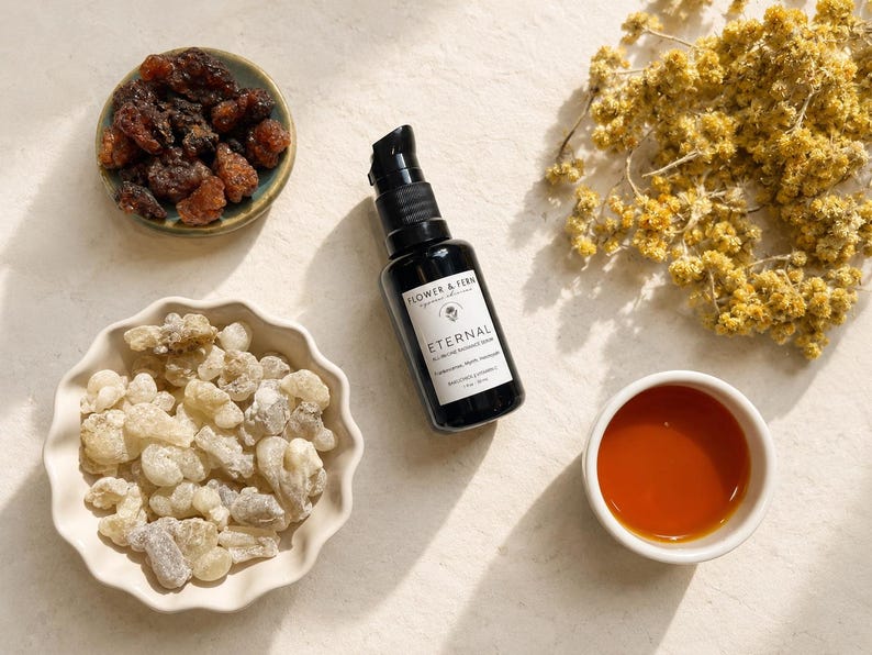 May include: A black pump bottle labeled Flower & Fern Eternal All-in-One Radiance Serum 1 fl oz / 30 ml, surrounded by a small bowl of dark resin, a scalloped bowl of light resin chunks, a small cup of amber liquid, and dried yellow flowers.
