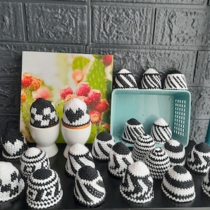 May include: A collection of black and white crocheted egg-shaped ornaments with various patterns, including hearts, stripes, and geometric designs. The ornaments are displayed on a table with a light blue background.
