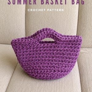 Może przedstawiać: Fioletowa szydełkowa torba koszykowa ze zintegrowanymi uchwytami. Tekst SUMMER BASKET BAG CROCHET PATTERN jest wyświetlany ciemnofioletowymi literami nad torbą. Torba ma grubą, teksturowaną konstrukcję z przędzy o zaokrąglonym, otwartym kształcie.