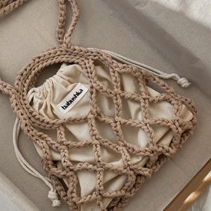 Può includere: Borsa tote in macramè beige con chiusura a coulisse. La borsa presenta un design a rete intrecciata e un sacchetto beige più piccolo all'interno. L'etichetta sulla borsa recita "babushka".