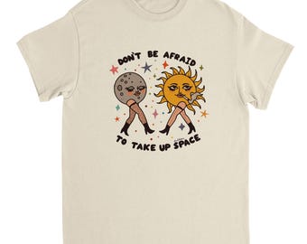 May include: A cream-coloured t-shirt with a retro illustration of the sun and moon, both with legs, fishnet stockings, and boots. The text "DON'T BE AFRAID TO TAKE UP SPACE" is printed above and below the figures in a vintage font.