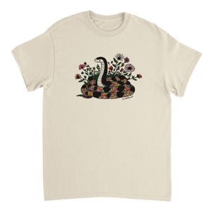Peut inclure: T-shirt crème avec un motif de serpent coloré. Le serpent est noir et blanc avec un motif arc-en-ciel sur son corps, entouré de fleurs. Le motif est centré sur le t-shirt.