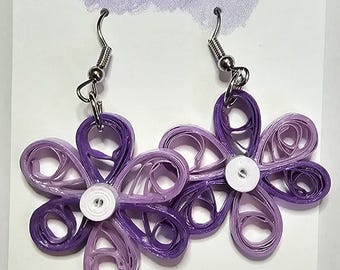 May include: A pair of handmade flower earrings in shades of purple, with a white center. The earrings are made of rolled paper and hang from silver-colored hooks. The background is a white card with the text "Artfully Loved" in purple.