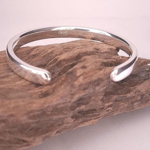 May include: A polished silver cuff bracelet with rounded ends. The bracelet is stamped with the number "(999)" and rests on a piece of driftwood. The open design allows for easy wear and removal. This minimalist bracelet is a versatile accessory.