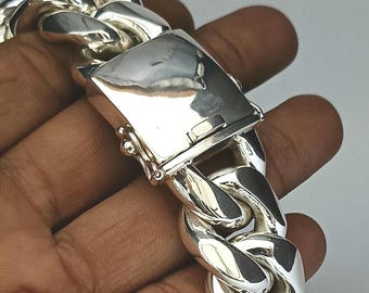 May include: A close-up of a silver chain bracelet with large, interlocking links. The bracelet features a rectangular clasp and a polished, reflective surface. The jewelry is displayed against a neutral background.