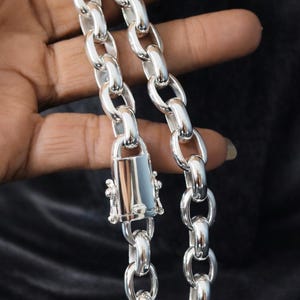 May include: Close-up of a silver chain necklace with large, oval links. The necklace features a decorative clasp with intricate detailing. The chain is displayed against a dark background, highlighting its shiny, metallic appearance.