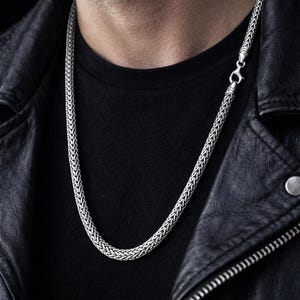 May include: A silver chain necklace with a woven design. The necklace is displayed against a black background, worn over a black shirt and a black leather jacket.