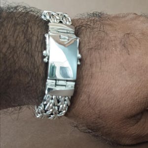 May include: A silver bracelet with a chain-link design and a rectangular clasp. The bracelet is made of a shiny, reflective metal. The clasp has a polished surface. The bracelet is worn on a wrist.