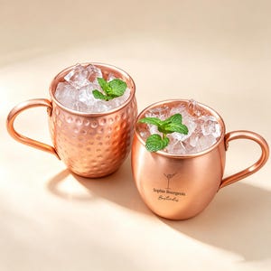 以下が含まれることがあります： 氷と新鮮なミントの葉が入った2つの銅製マグカップ。片方はハンマーで叩いたような槌目模様で、もう片方は滑らかな表面にマティーニグラスのアイコンと「Sophie Bourgeois Bartender」という黒い文字がプリントされています。